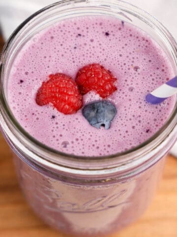 A glass jar filled with a creamy purple triple berry smoothie, topped with fresh raspberries and a blueberry, served with a purple-striped paper straw on a wooden surface.