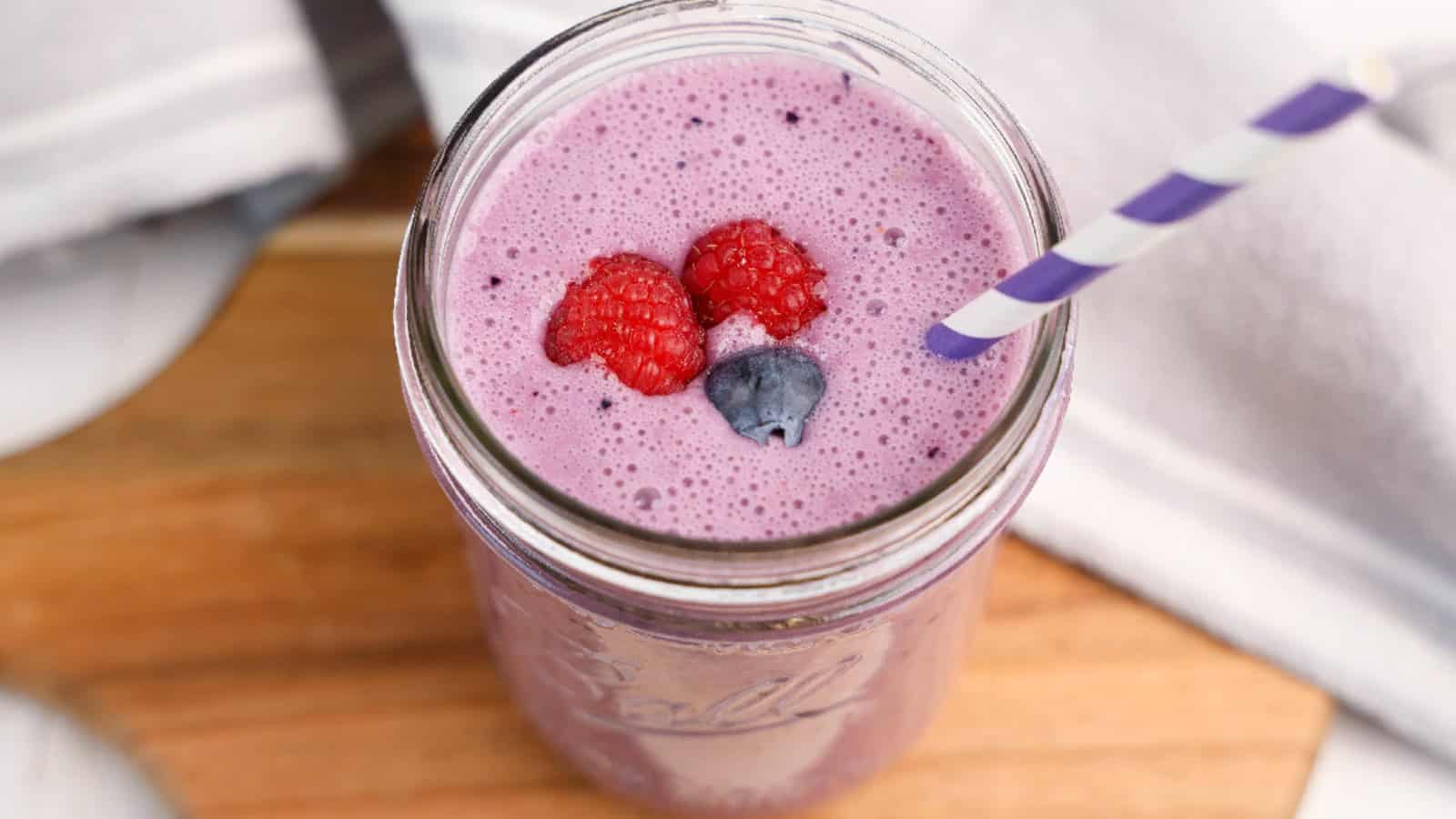A glass jar filled with a creamy purple triple berry smoothie, topped with fresh raspberries and a blueberry, served with a purple-striped paper straw on a wooden surface.