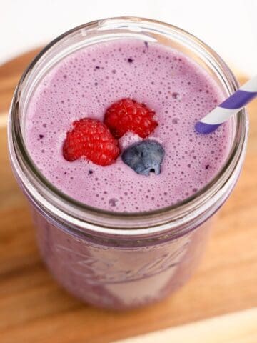 A glass jar filled with a creamy purple triple berry smoothie with a purple-striped paper straw.