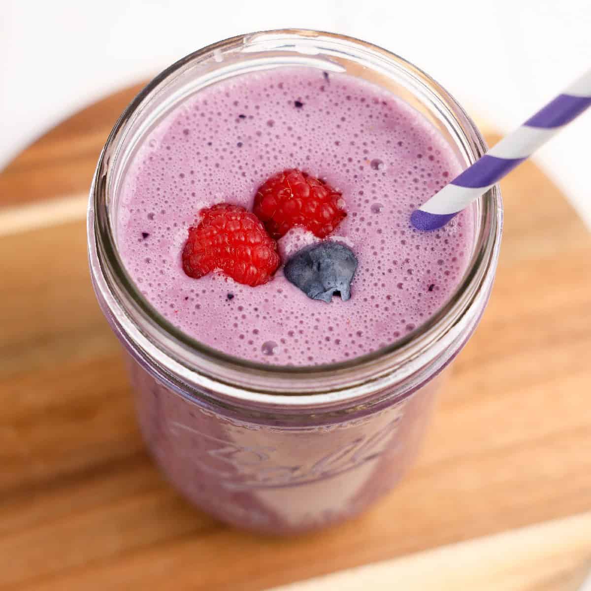A glass jar filled with a creamy purple triple berry smoothie with a purple-striped paper straw.