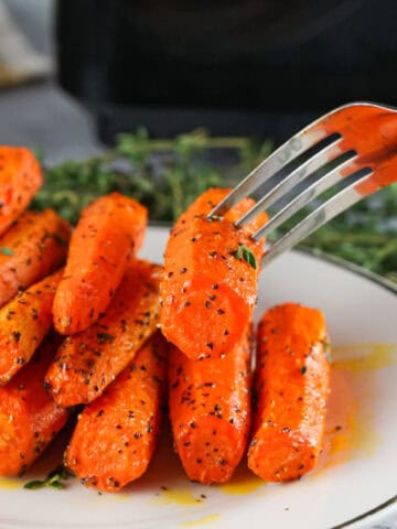Fork lifting a roasted air fryer carrot from a plate of herb-seasoned carrots.