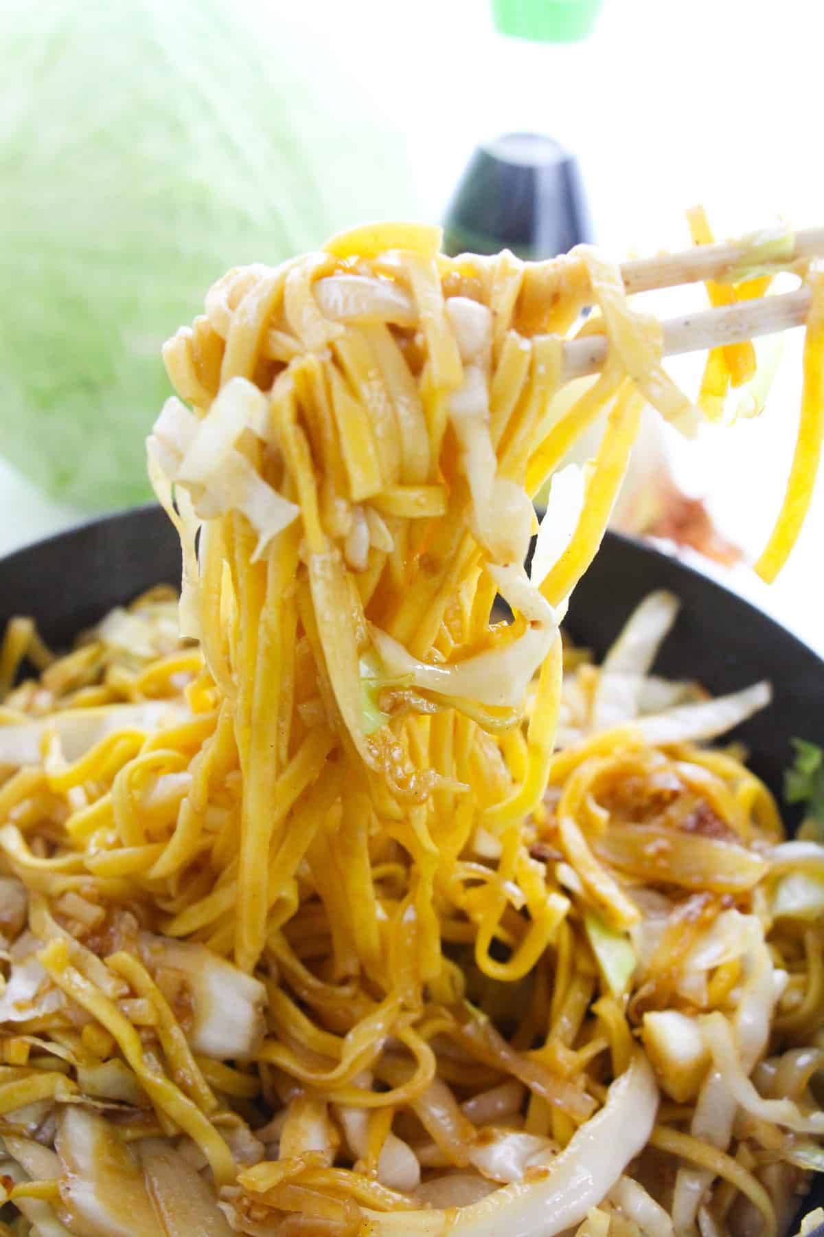 Chopsticks lifting cabbage chow mein noodles from a wok.
