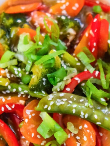 Veggie teriyaki stir fry with broccoli, carrots, red bell peppers, and snap peas in a savory sauce, topped with sesame seeds and green onions in a white bowl.