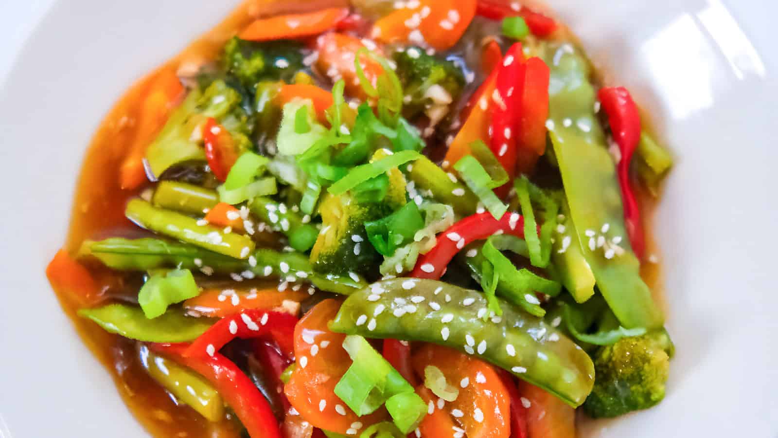 Veggie teriyaki stir fry with broccoli, carrots, red bell peppers, and snap peas in a savory sauce, topped with sesame seeds and green onions in a white bowl.