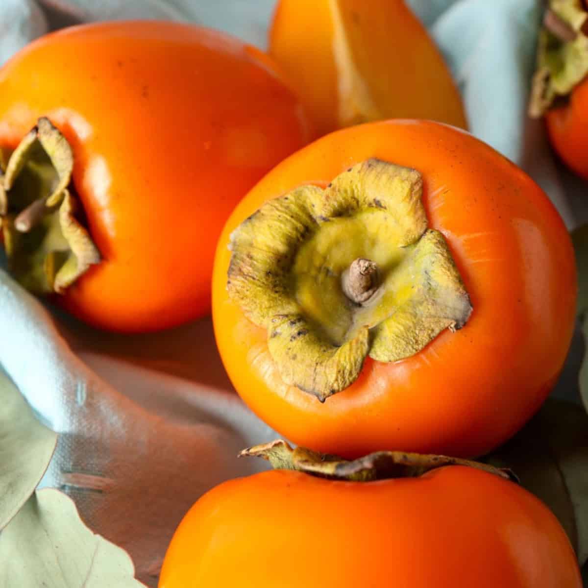 Fresh persimmons with vibrant orange skin and leafy tops resting on a light blue cloth.