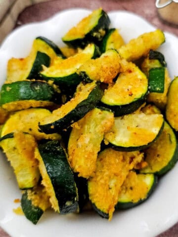 A white bowl filled with air-fried zucchini coated in vegan parmesan and breadcrumbs, placed on a rustic wooden surface with a plaid cloth in the background.