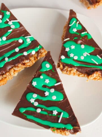 Close-up of three Christmas tree-shaped peanut butter bars on a white plate, topped with melted chocolate, green icing drizzle, and white sprinkles, creating a festive holiday dessert.