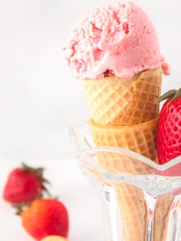 Vegan strawberry ice cream scooped into a waffle cone, served in a glass cup with fresh strawberries around it.