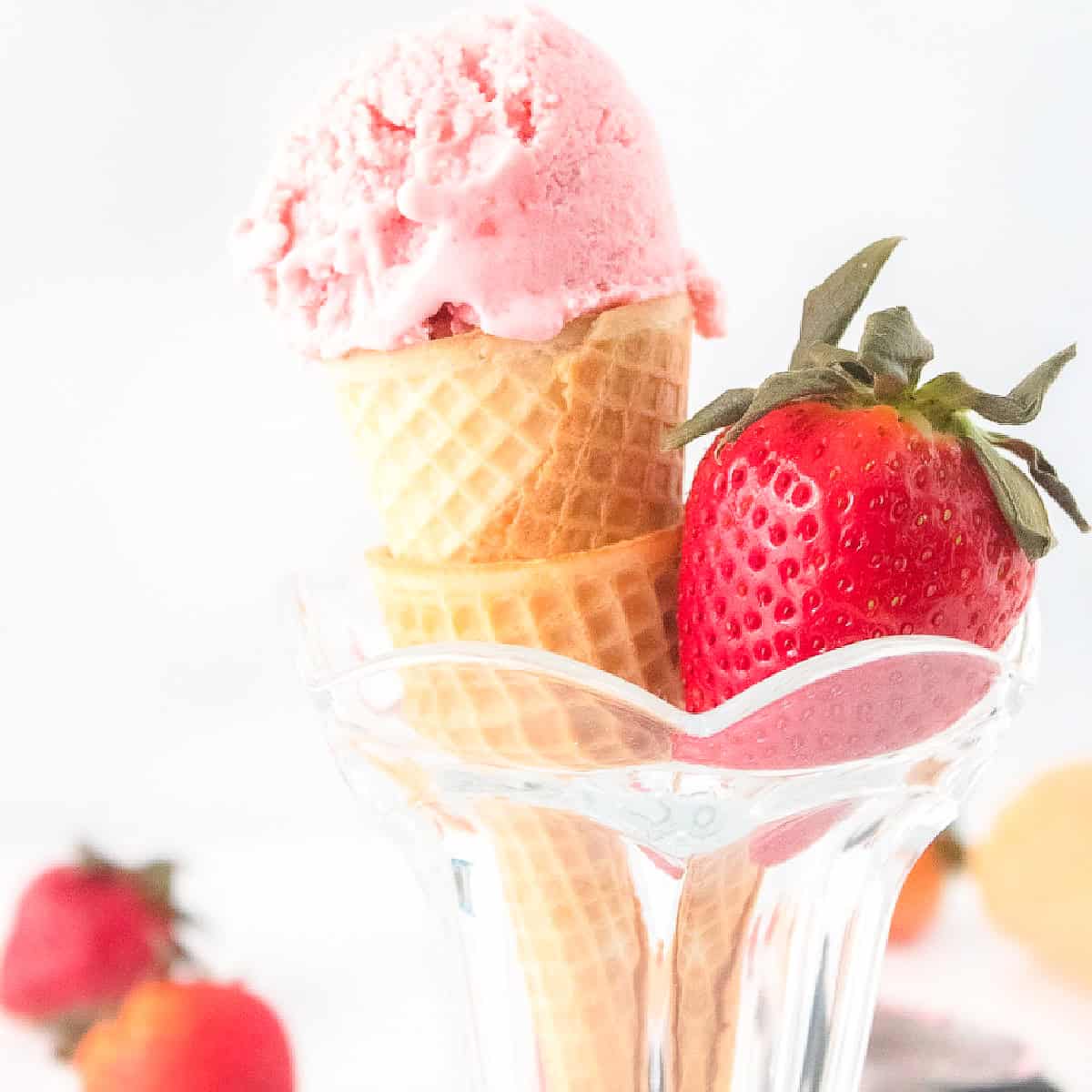 A scoop of creamy pink strawberry ice cream sits on a waffle cone in a glass cup, next to a fresh ripe strawberry.
