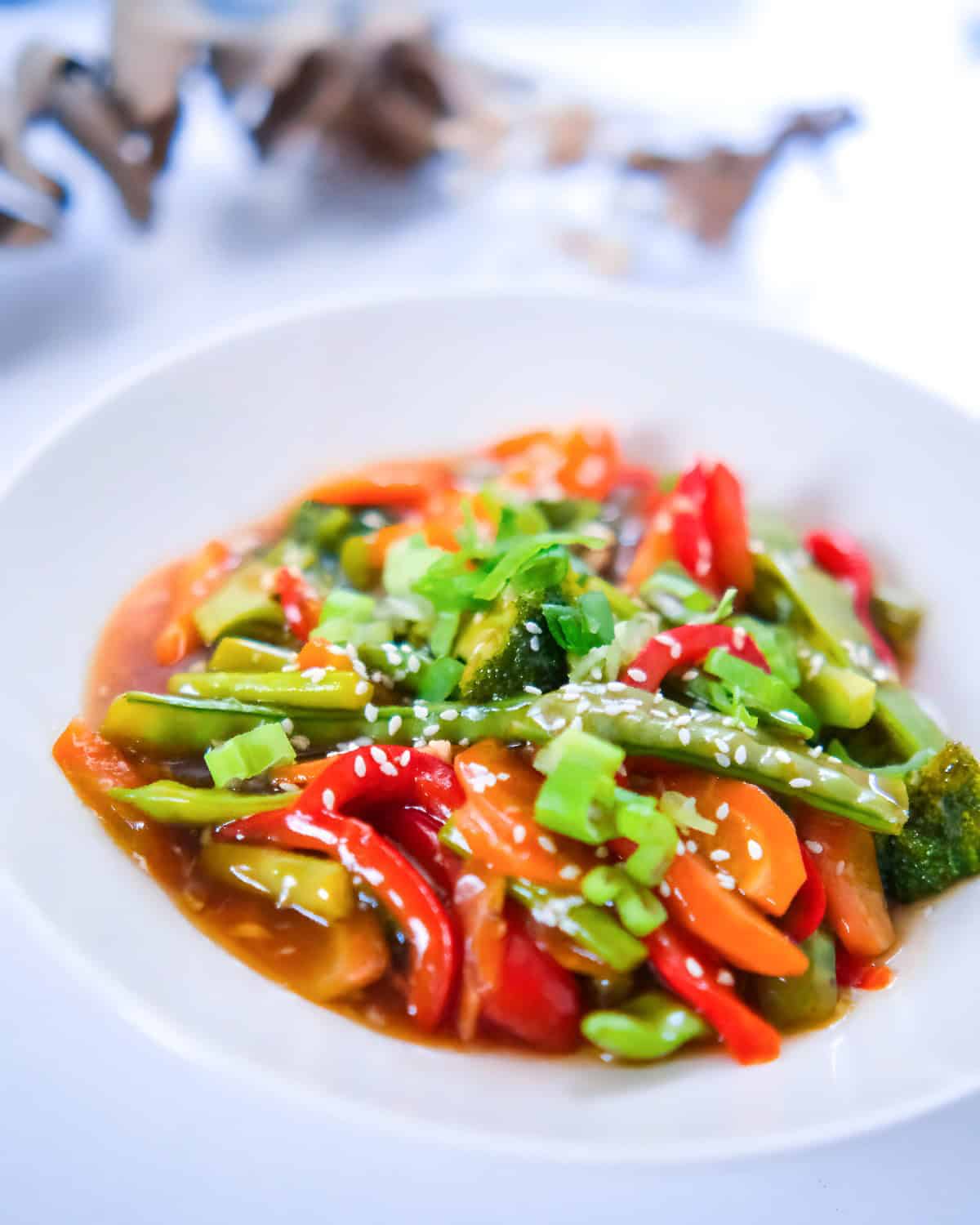 Closer view of teriyaki stir fry vegetables in a white bowl, topped with sesame seeds and fresh scallions for garnish.
