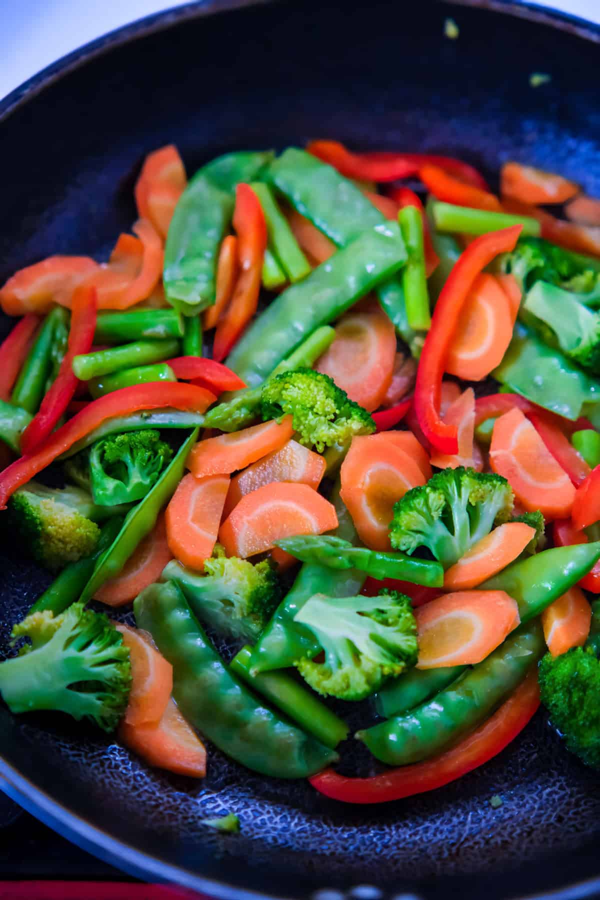 Fresh vegetables including broccoli, carrots, red bell peppers, and snow peas being stir-fried in a pan.
