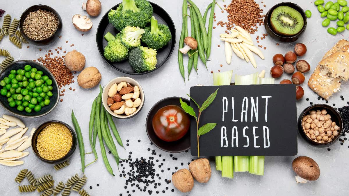 An assortment of plant-based foods such as nuts, seeds, beans, broccoli, and vegetables arranged around a sign that reads “plant based.”