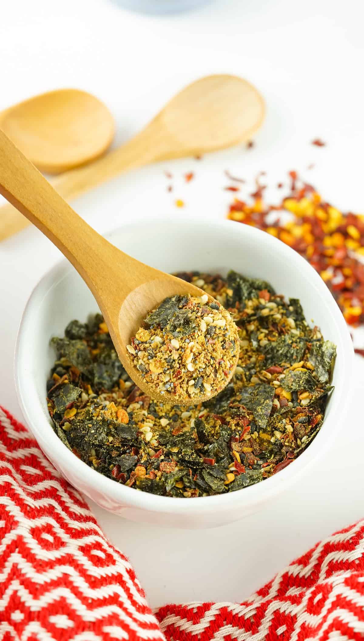 Wooden spoon scooping homemade Japanese 7 spice seasoning from a white bowl with seaweed, sesame seeds, and chili flakes.