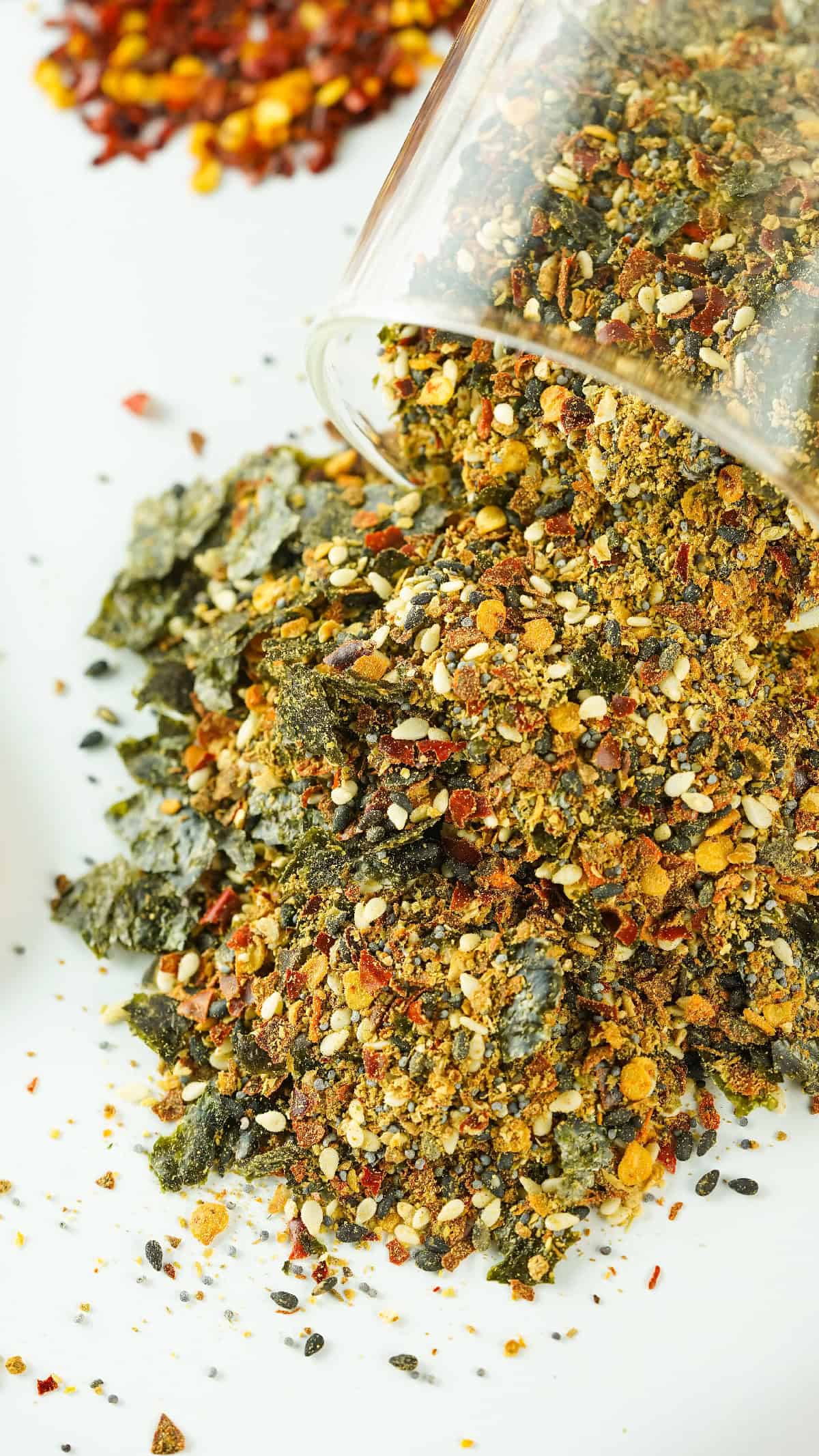 Close-up of Japanese 7 spice seasoning spilling out of a glass jar, showing seaweed flakes, sesame seeds, and chili flakes.