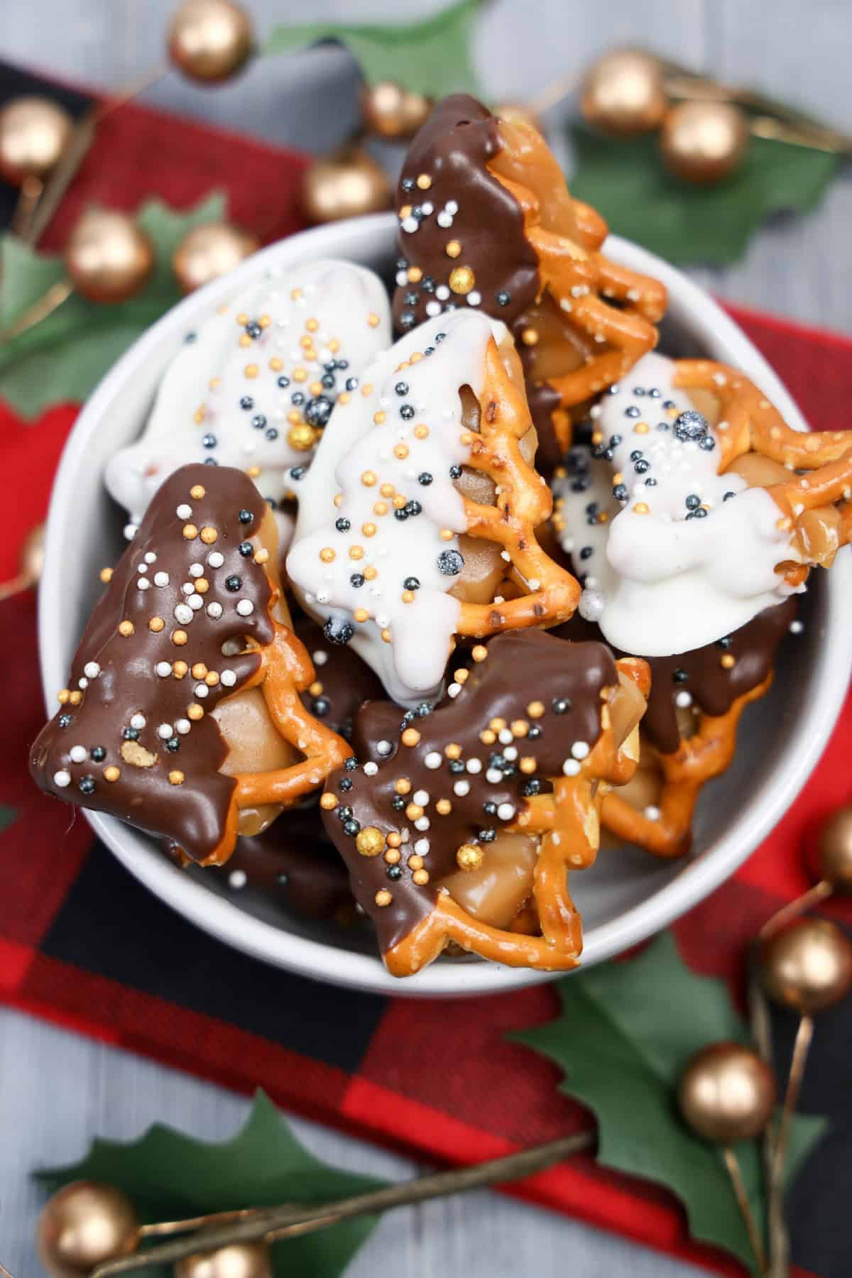 Bowl of caramel-filled holiday pretzels dipped in white and dark chocolate with festive sprinkles.