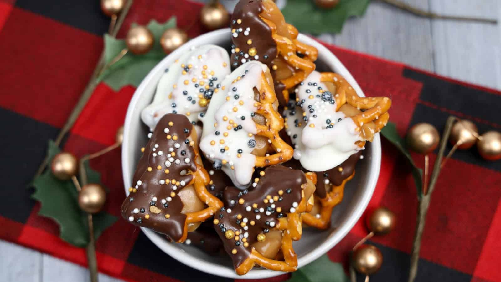 Bowl of New Year’s Eve dipped caramel pretzels shaped like holiday trees, covered in white and dark chocolate with festive gold and silver sprinkles, on a red plaid background.