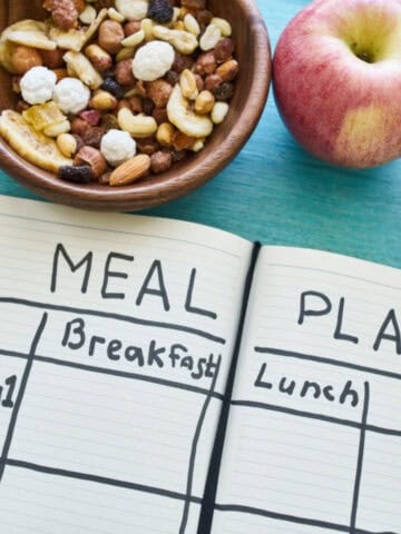 Open meal planning notebook with sections for breakfast, lunch, and dinner next to a bowl of mixed nuts and fresh apples on a table.