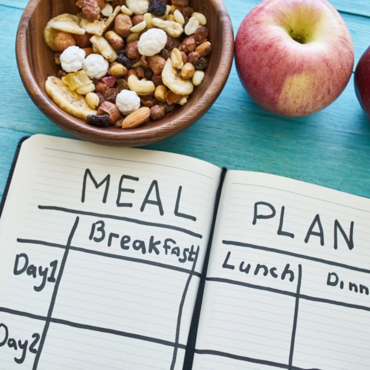 Open meal planning notebook with sections for breakfast, lunch, and dinner next to a bowl of mixed nuts and fresh apples on a table.