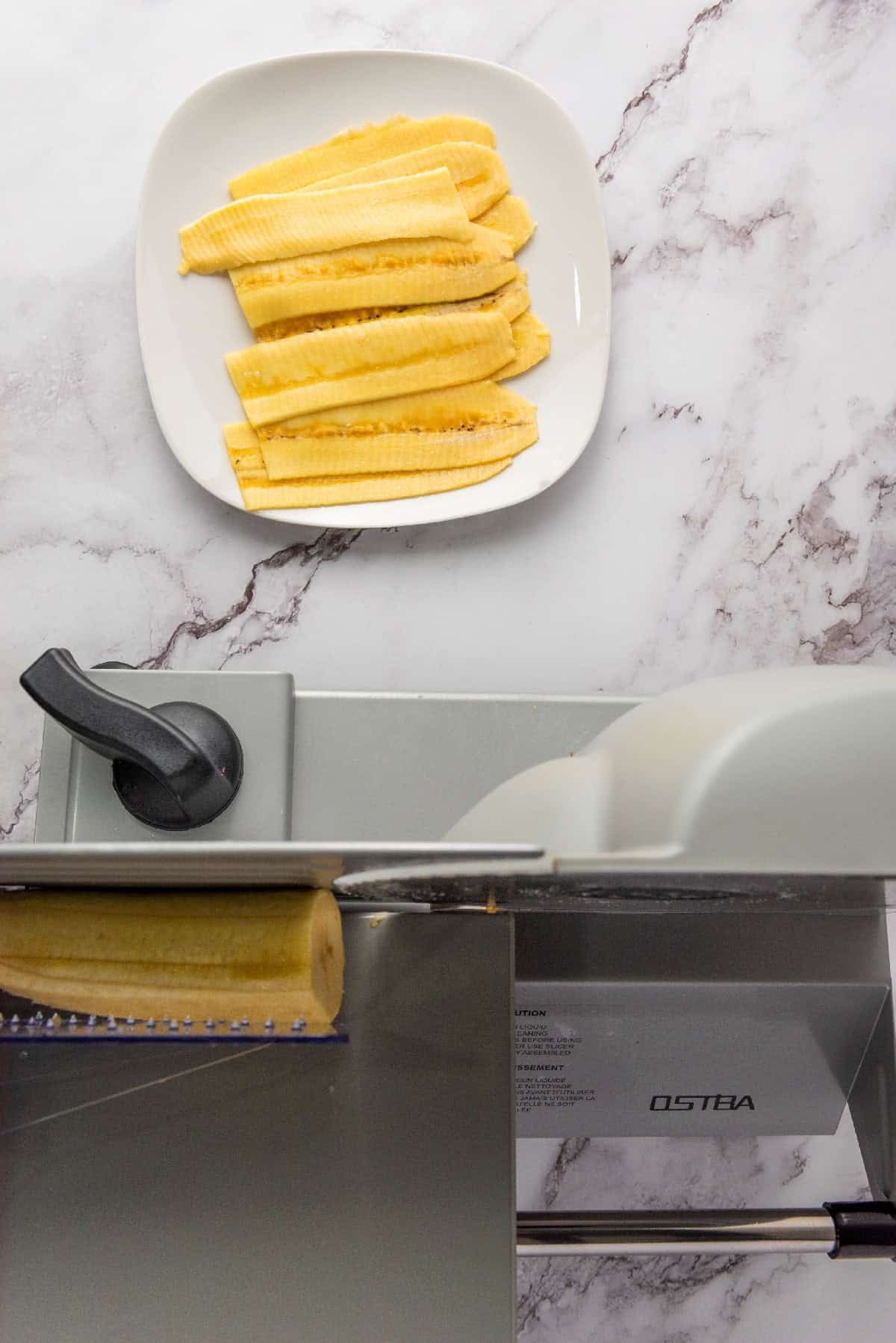 Thinly sliced plantains arranged on a plate next to a mandoline slicer.