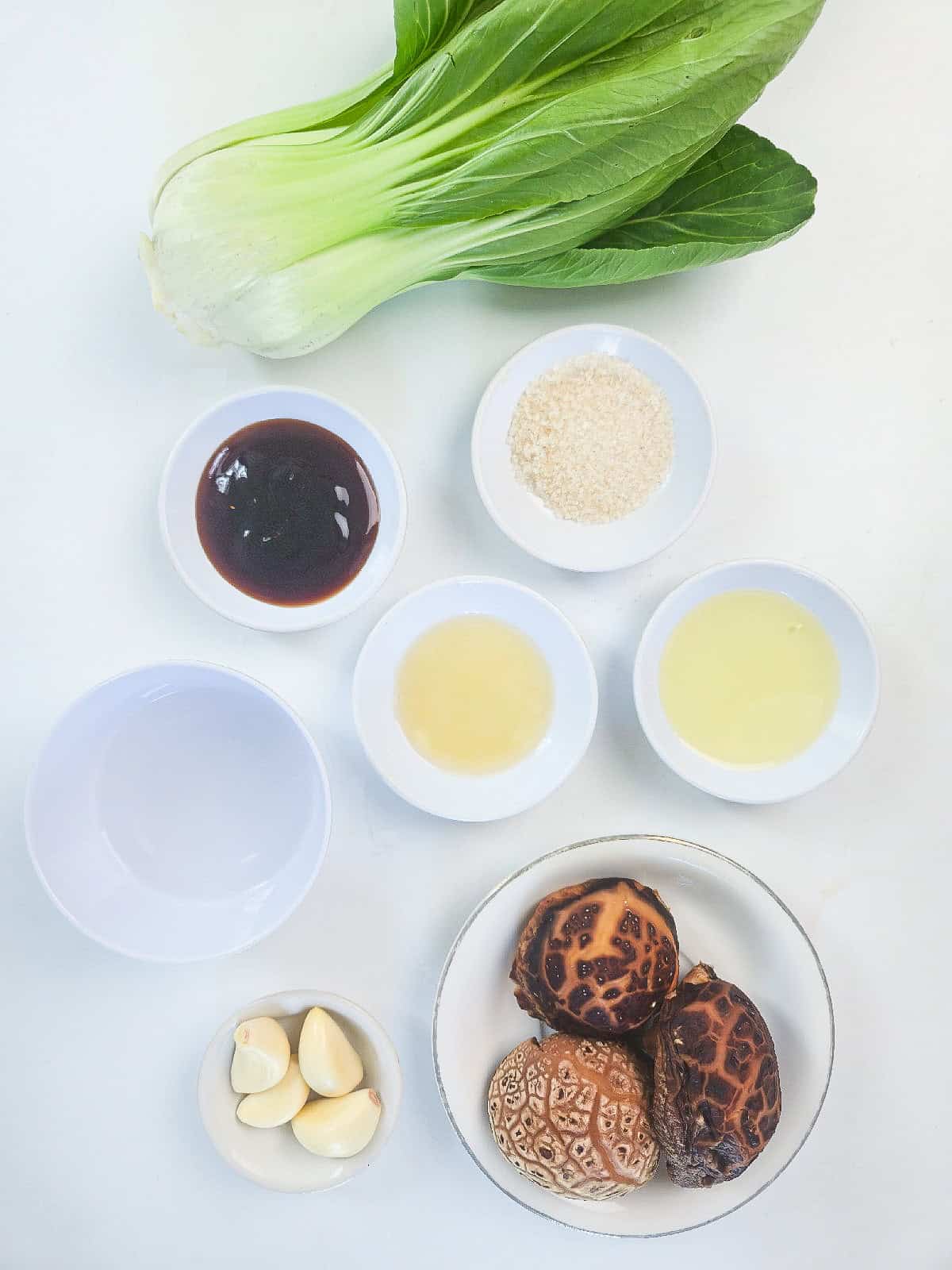 Bok choy, dried shiitake mushrooms, garlic, and stir-fry sauce ingredients arranged on a white surface.