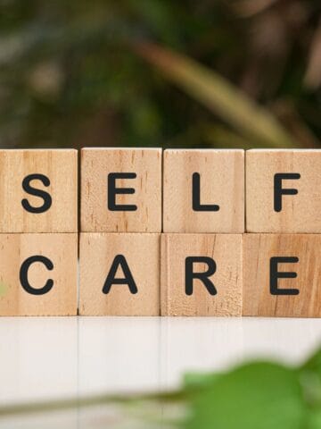 Wooden letter blocks spelling “self care” on a table surrounded by green plants.