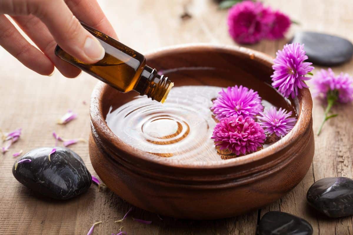 A hand pours essential oil from a small bottle into a wooden bowl of water with floating pink flowers.