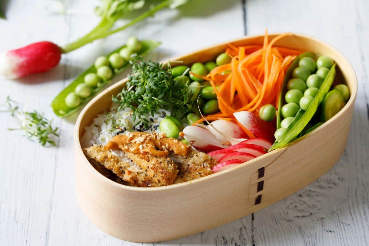 Wooden bento lunch box with rice, breaded fish, edamame, shredded carrots, radishes, and fresh greens.