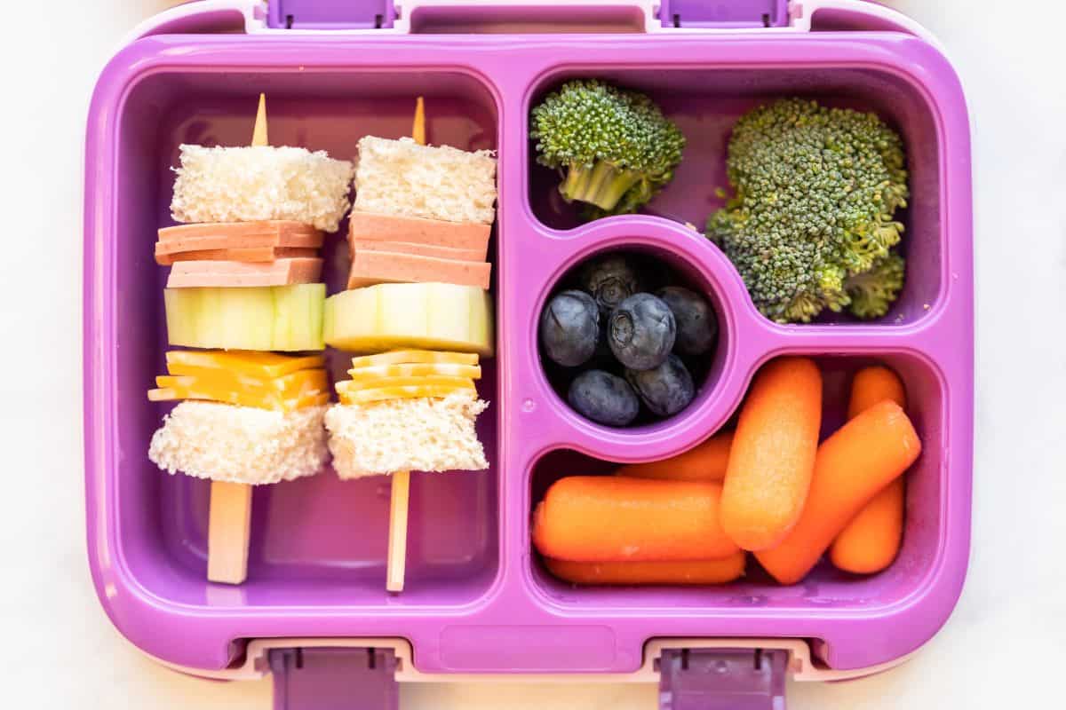 A colorful bento lunch box with sandwich skewers, broccoli, blueberries, and baby carrots in separate compartments.