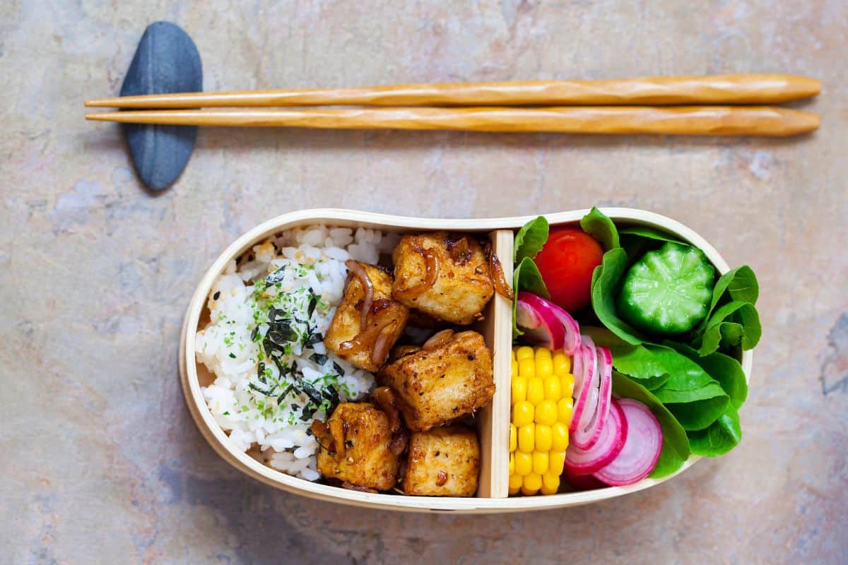 Wooden bento lunch box with rice topped with seaweed, glazed tofu cubes, corn, cucumber, tomato, pickled onions, and greens, with chopsticks above.
