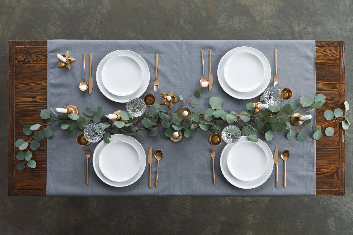 Overhead view of a neatly arranged dining table set with white plates, gold cutlery, candles, and greenery.