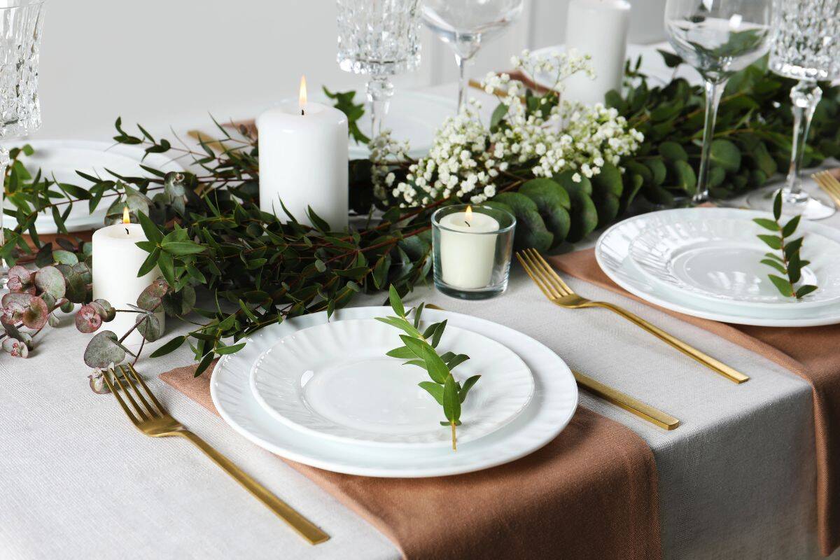 Elegant table setting with white plates, gold cutlery, candles, and greenery centerpiece.