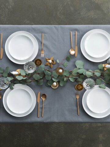 Top-down view of a simple table arrangement featuring white plates, neatly folded napkins, candles, and a greenery centerpiece.