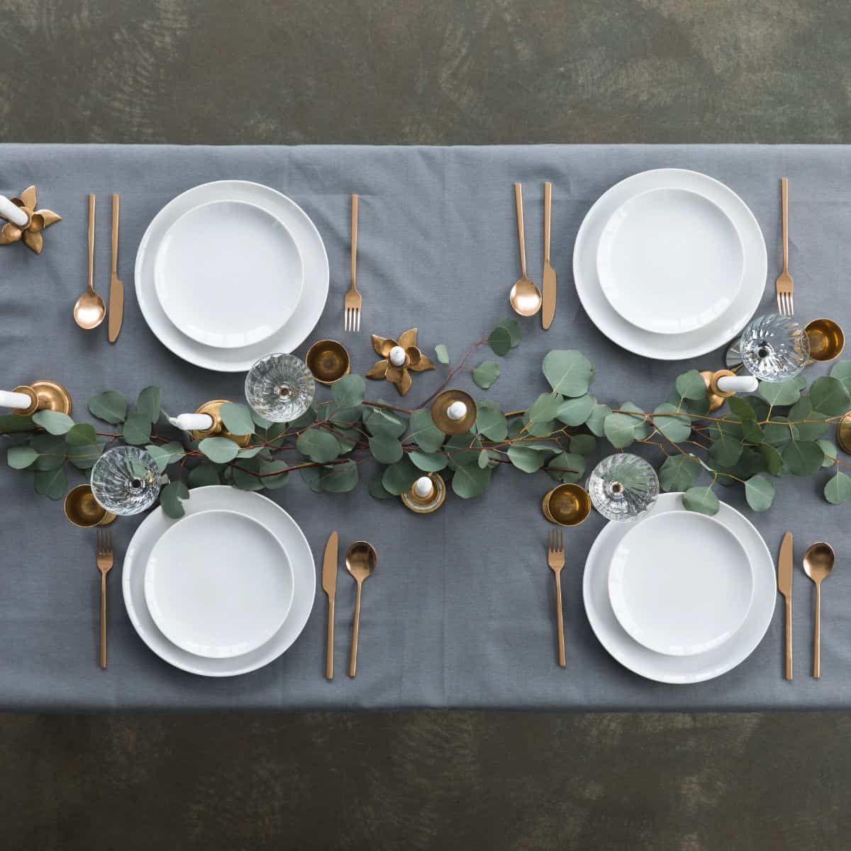 Top-down view of a simple table arrangement featuring white plates, neatly folded napkins, candles, and a greenery centerpiece.
