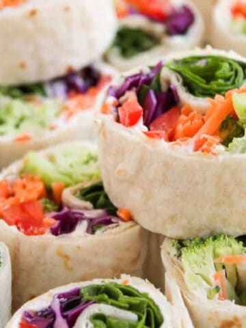 Colorful vegetable wrap pinwheels with broccoli, carrots, red cabbage, and spinach stacked on a white plate.