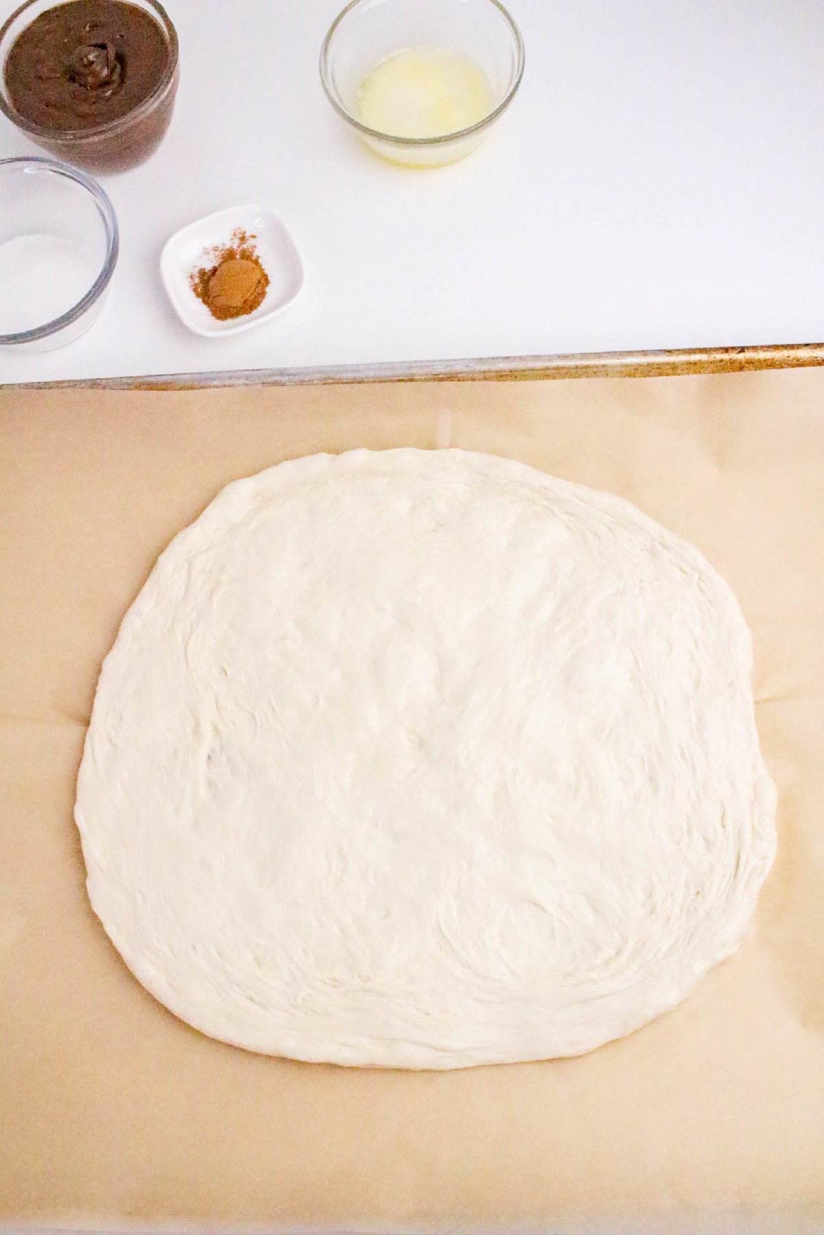 Pizza dough stretched on parchment paper with Nutella, butter, sugar, and cinnamon ingredients arranged above for Nutella dessert pizza.