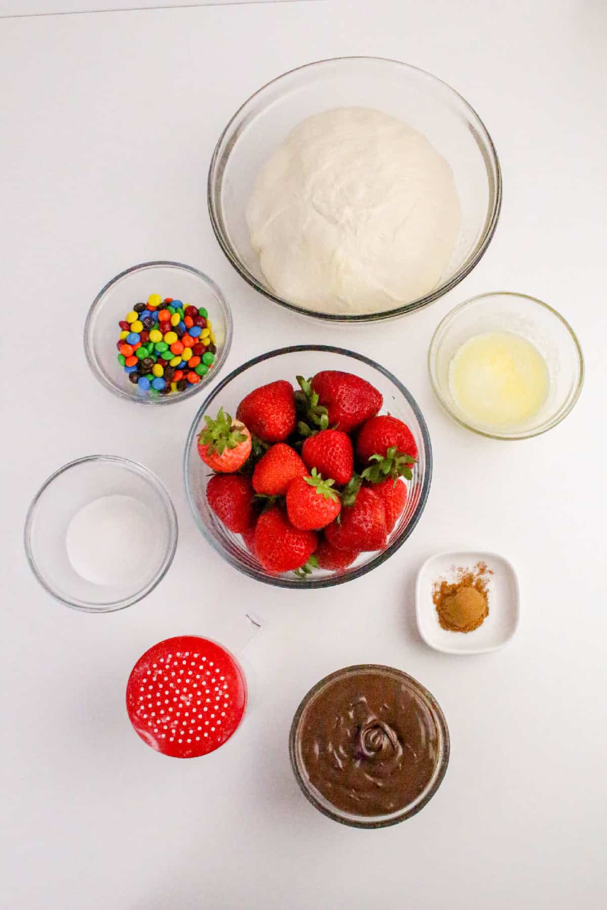 Ingredients for Nutella dessert pizza including pizza dough, fresh strawberries, Nutella, cinnamon sugar, butter, powdered sugar, and colorful candy toppings.