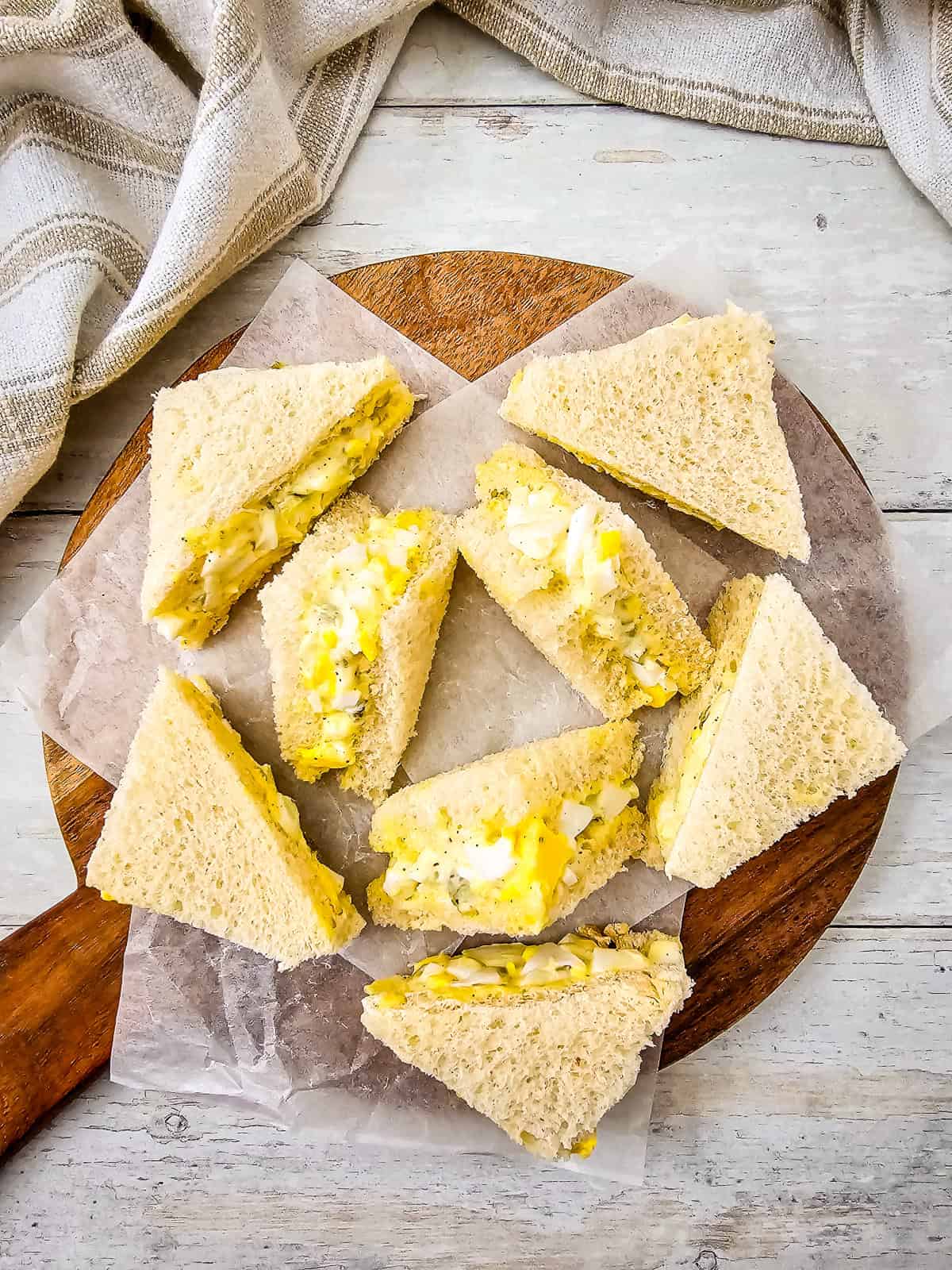 Egg salad tea sandwiches arranged on a wooden board with parchment paper, showing soft white bread and creamy egg filling.