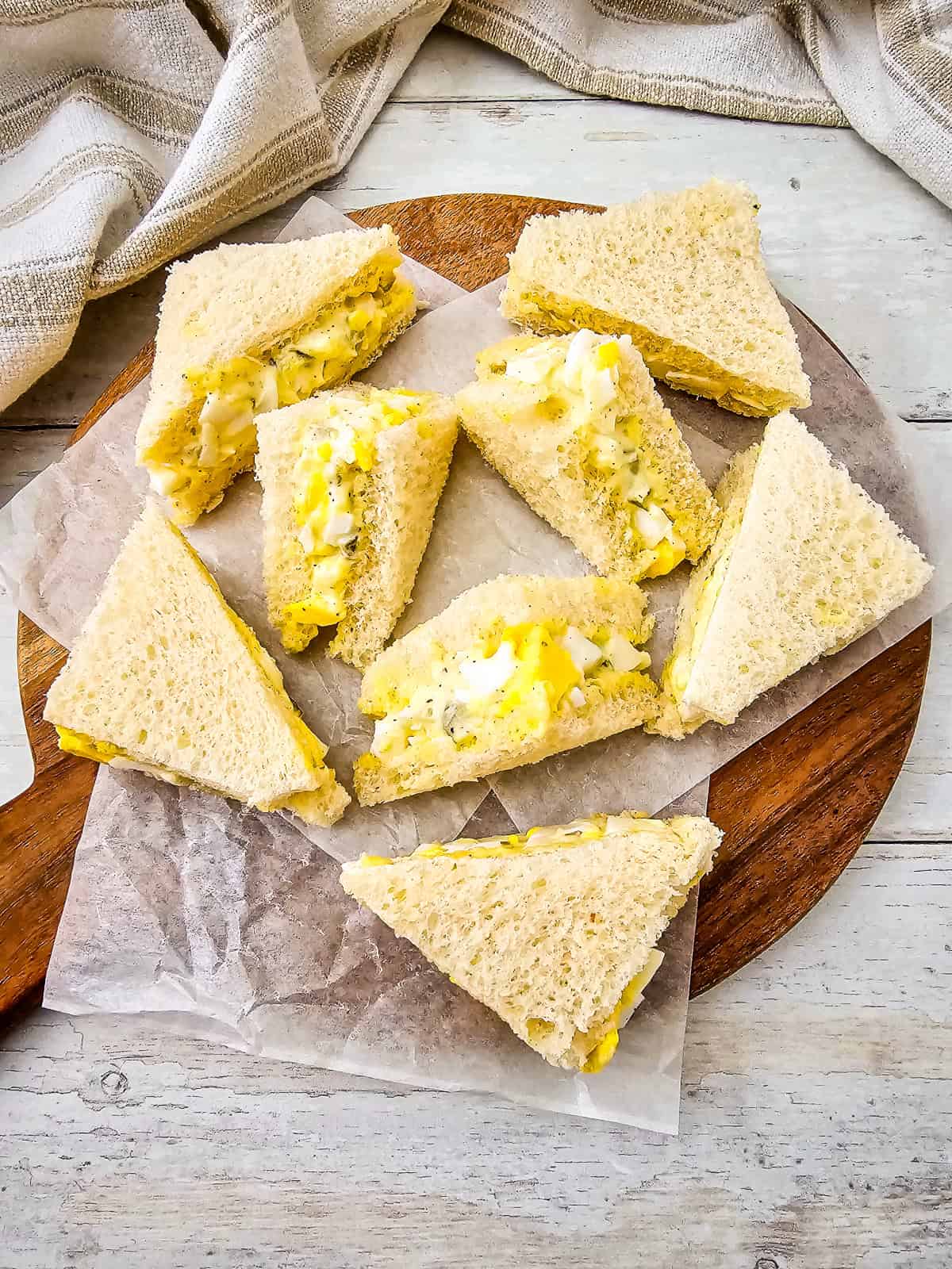Homemade egg salad tea sandwiches served on a wooden board with rustic cloth background.