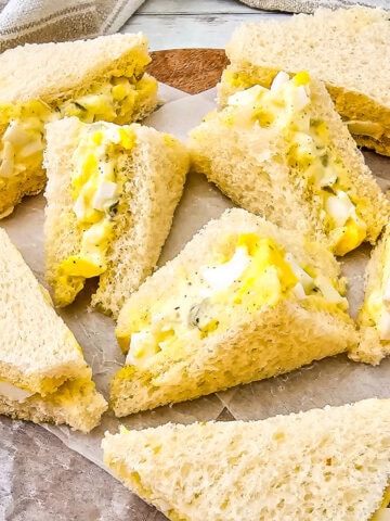 Egg salad tea sandwiches arranged on a wooden board with parchment paper, served with soft white bread and creamy filling.
