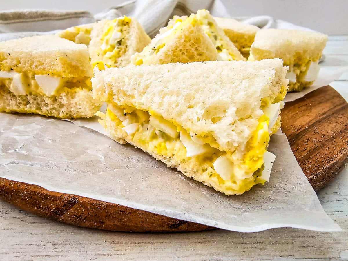 Homemade egg salad tea sandwiches on a wooden board with soft bread and rich egg filling.