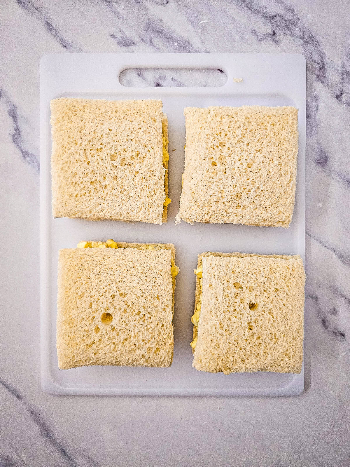 Egg salad tea sandwiches assembled with top bread slices on a cutting board.