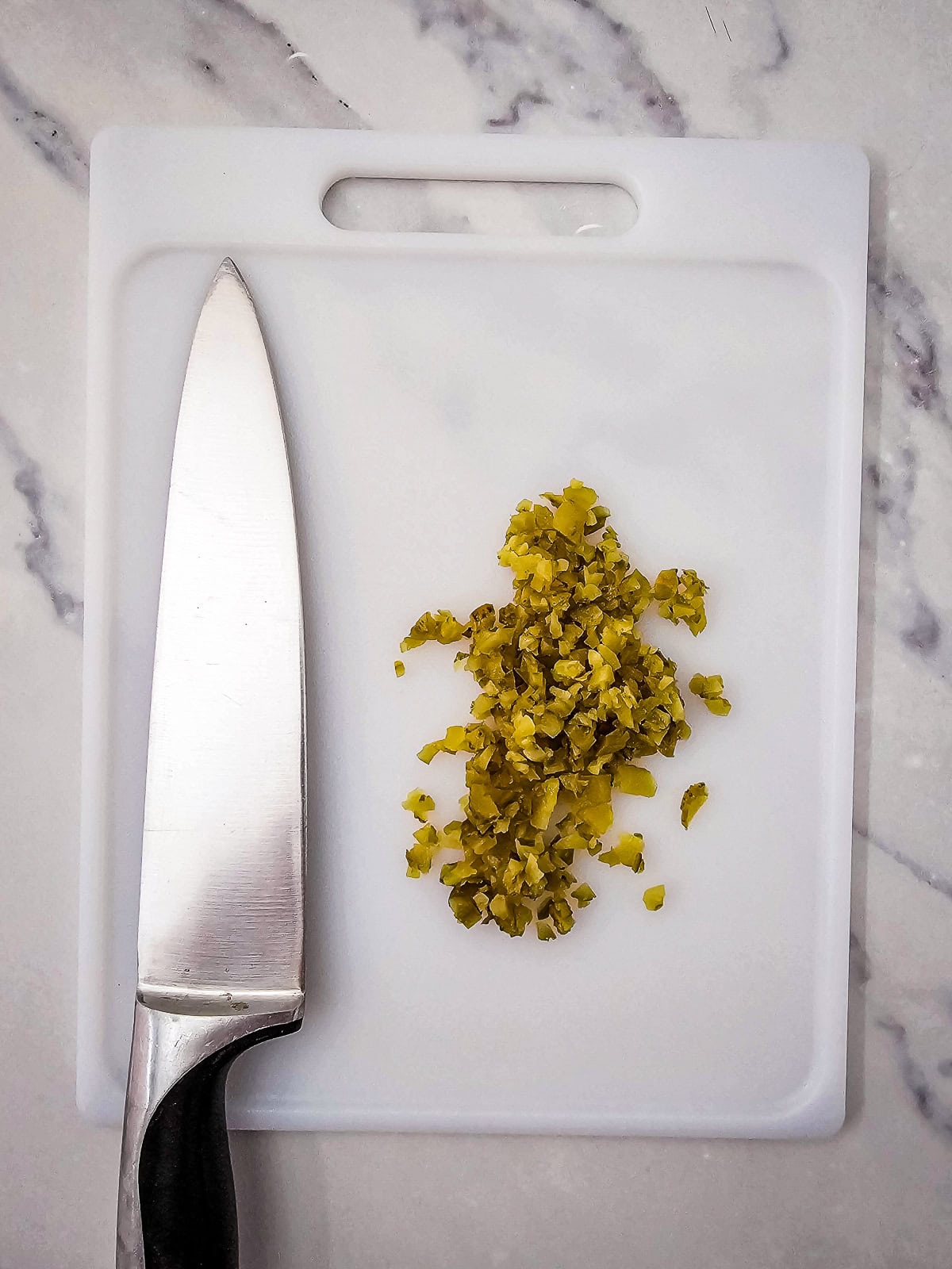Finely chopped dill pickles on a white cutting board with a chef's knife.