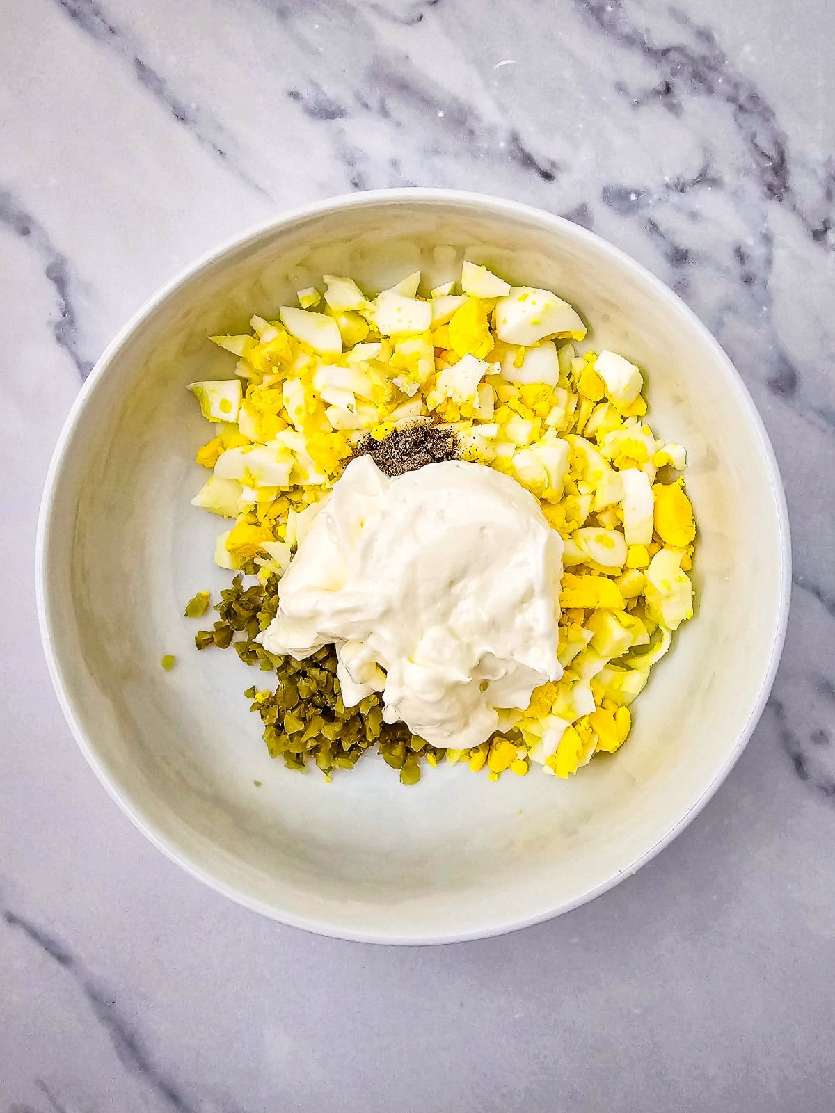 Egg salad ingredients with chopped eggs, pickles, black pepper, and mayonnaise in a mixing bowl.