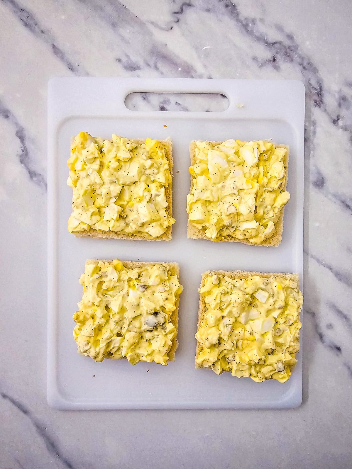 Egg salad spread evenly on small square bread slices for tea sandwiches.