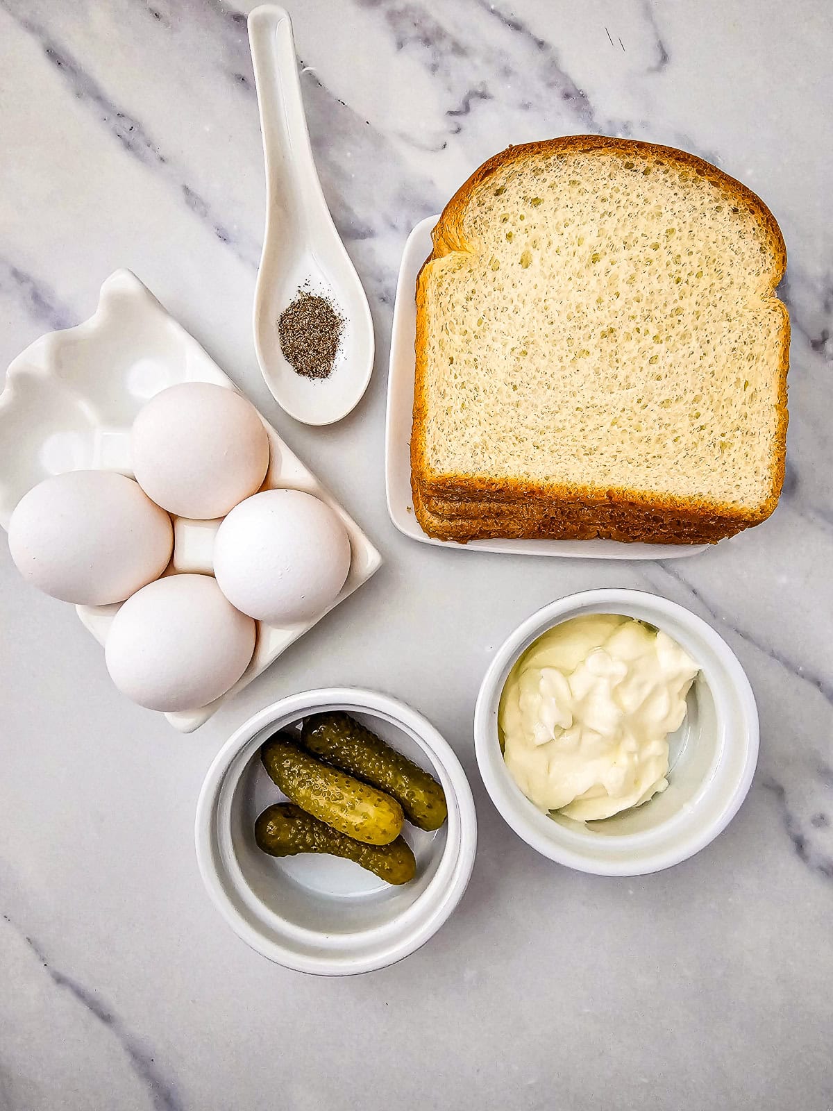 Ingredients for egg salad tea sandwiches including eggs, white bread, mayonnaise, pickles, and black pepper on marble surface.