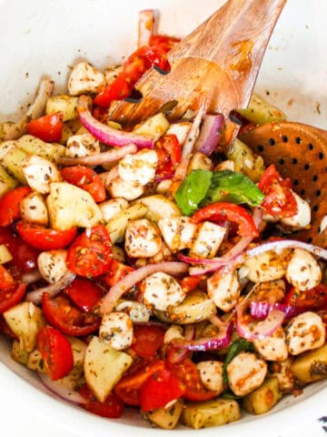 Cucumber caprese salad tossed with cherry tomatoes, vegan mozzarella pearls, red onion, fresh basil, and herb dressing in a white serving bowl.