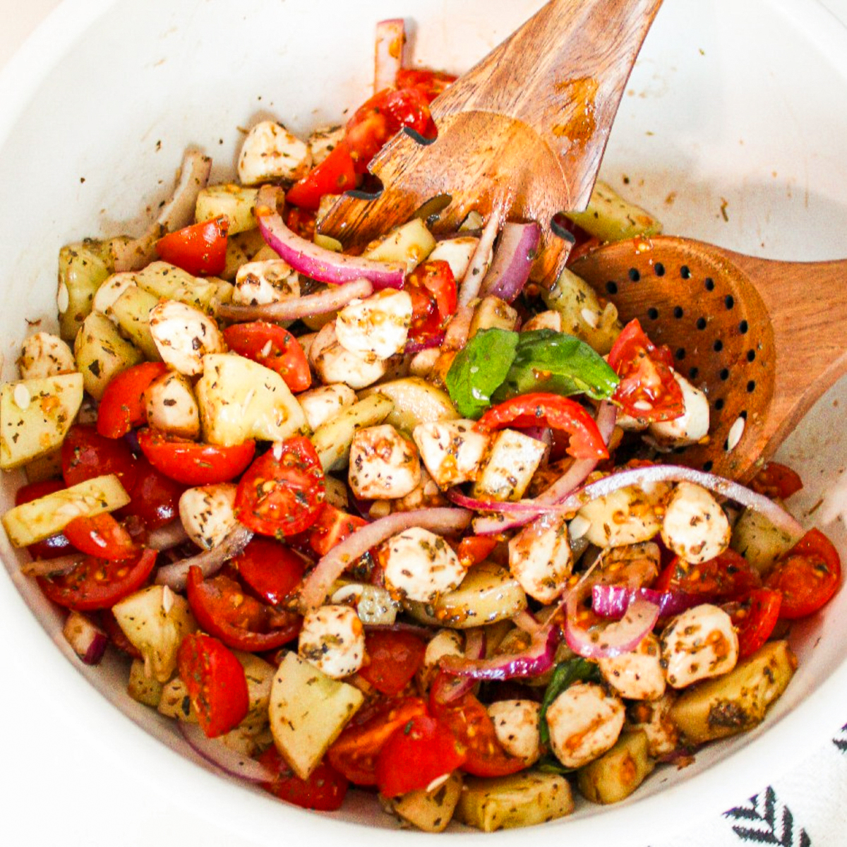 Cucumber caprese salad tossed with cherry tomatoes, vegan mozzarella pearls, red onion, fresh basil, and herb dressing in a white serving bowl.