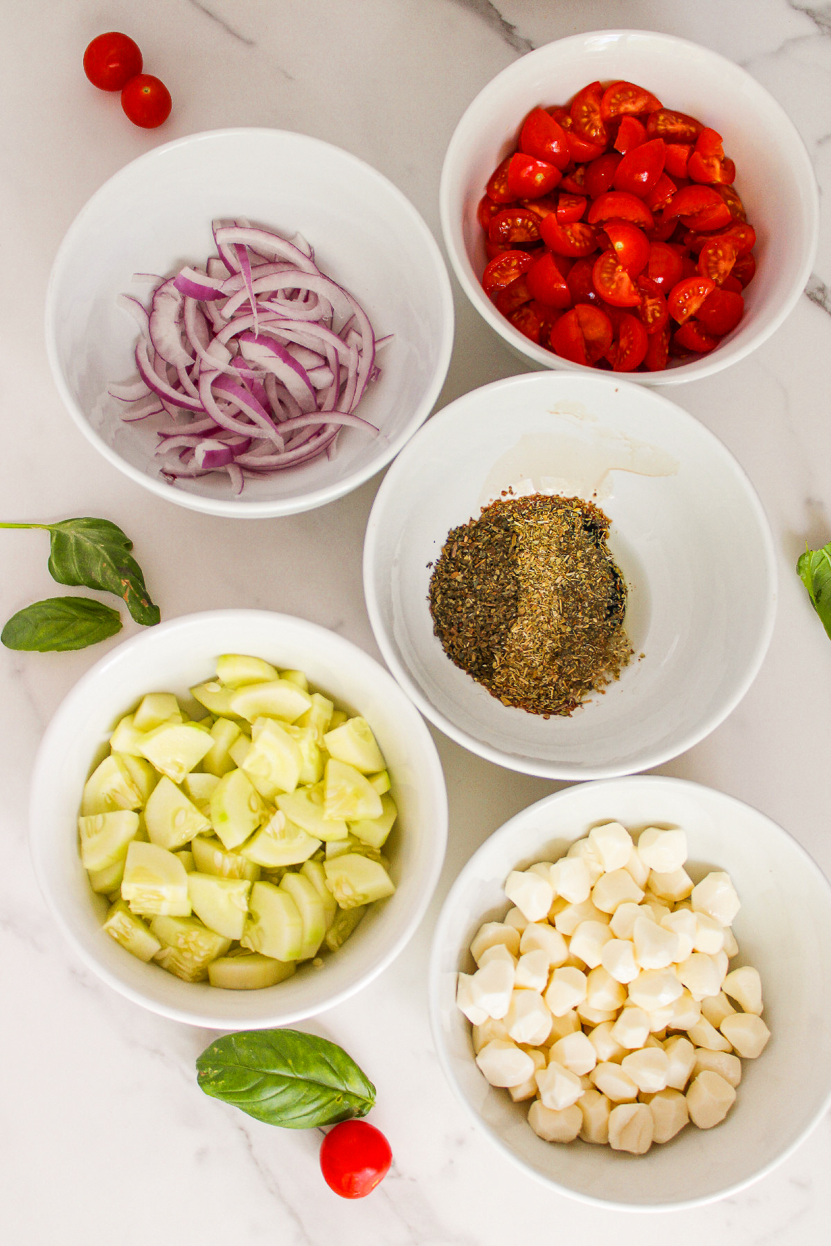 Ingredients for cucumber caprese salad arranged in white bowls, including chopped cucumber, cherry tomatoes, red onion, vegan mozzarella, fresh basil, and dried herbs on a marble countertop.