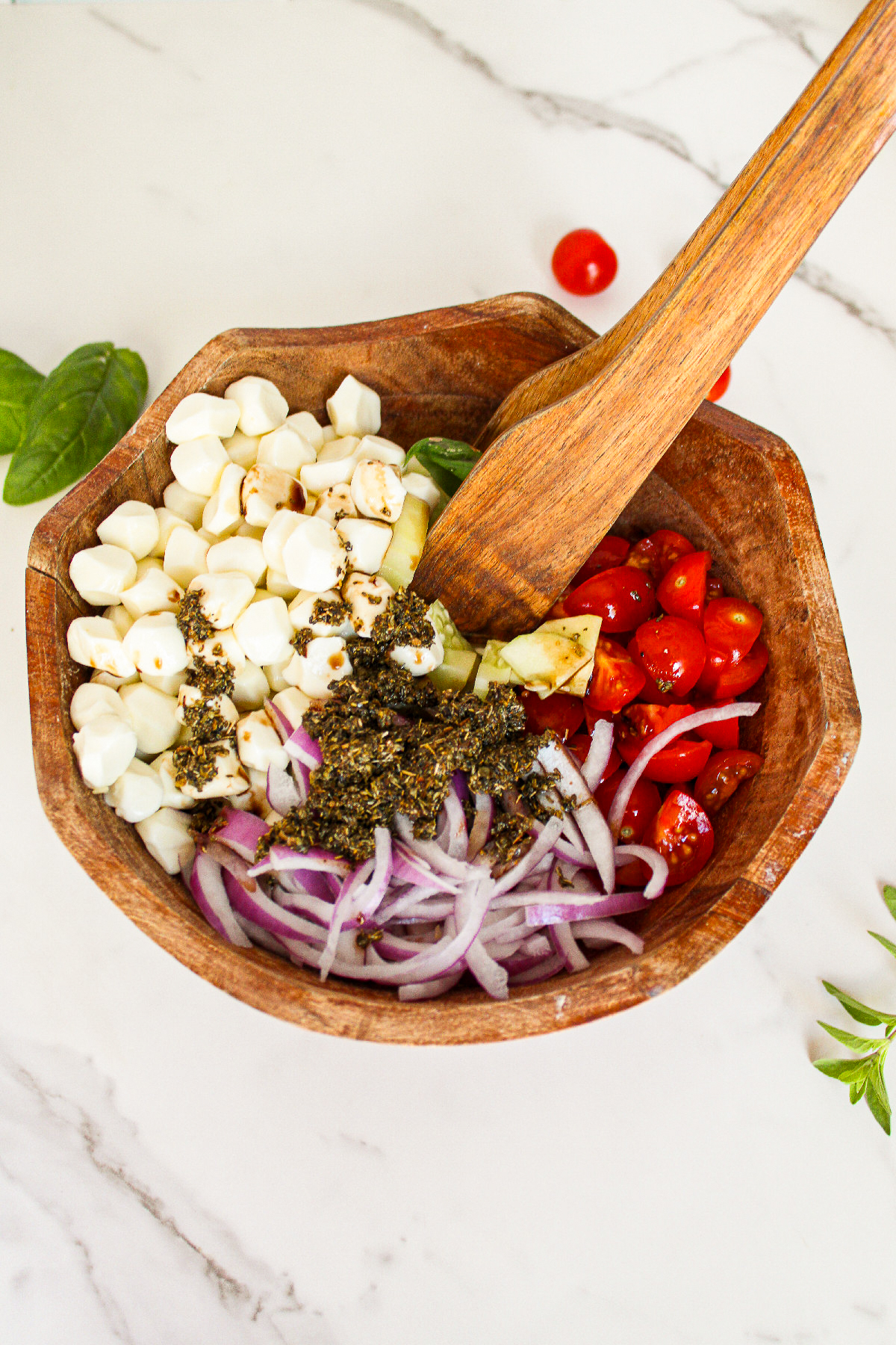 Herb dressing being added to cucumber caprese salad ingredients in a wooden serving bowl before tossing.