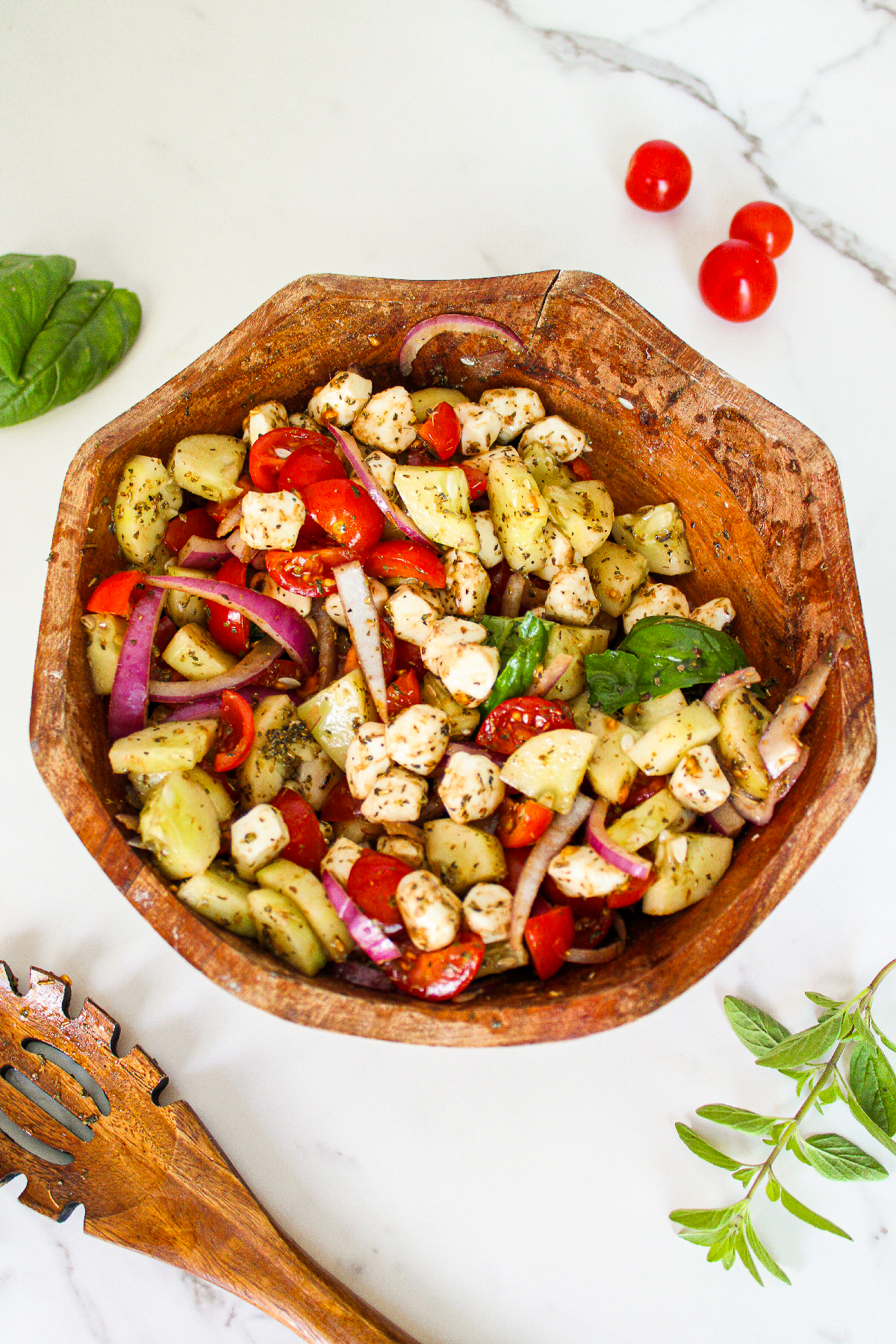 Cucumber caprese salad tossed with balsamic herb dressing, featuring cucumbers, cherry tomatoes, vegan mozzarella, red onion, and fresh basil in a wooden bowl.
